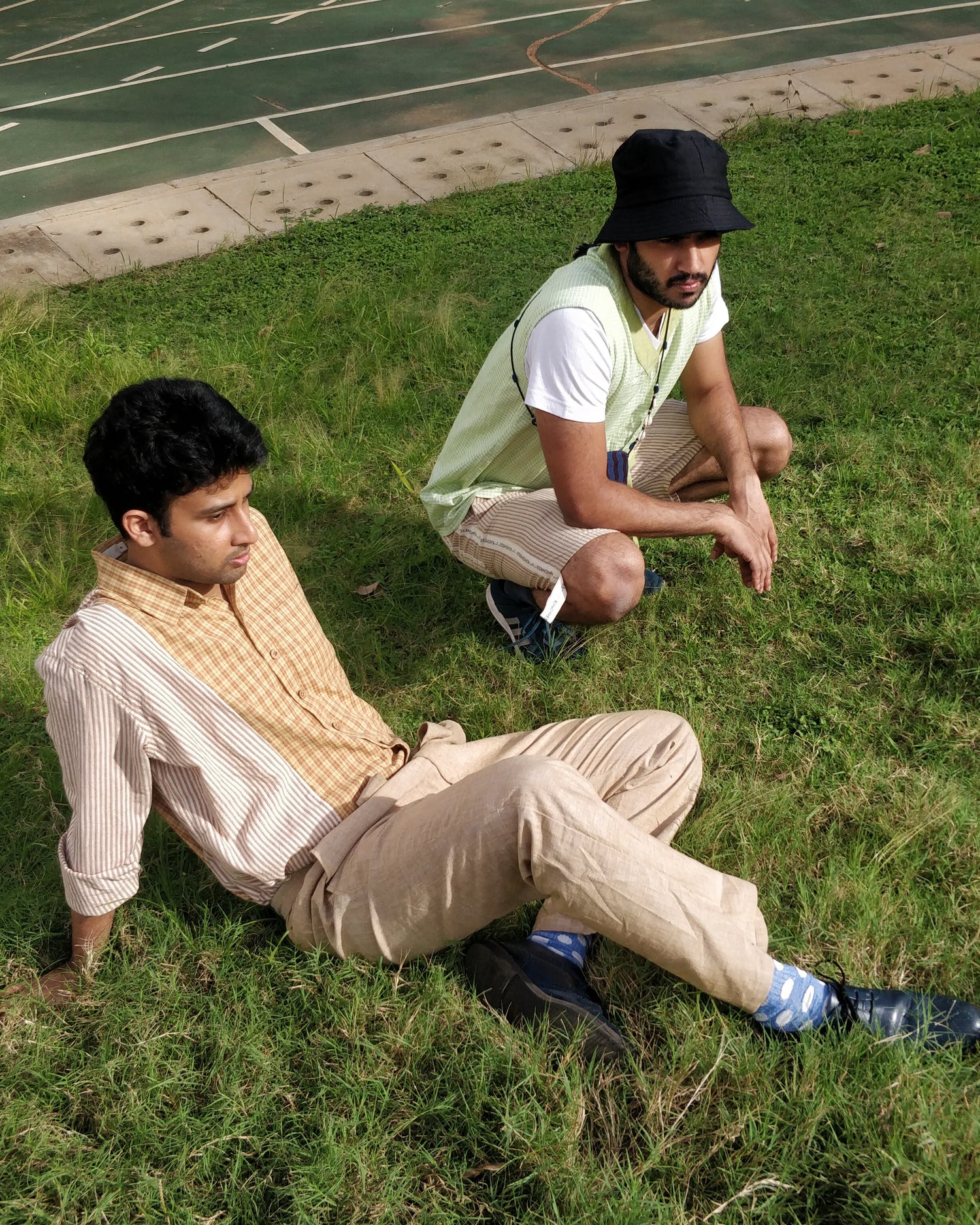 Two young men sitting on grass in comfortable clothes by KiRu. Checks and brown