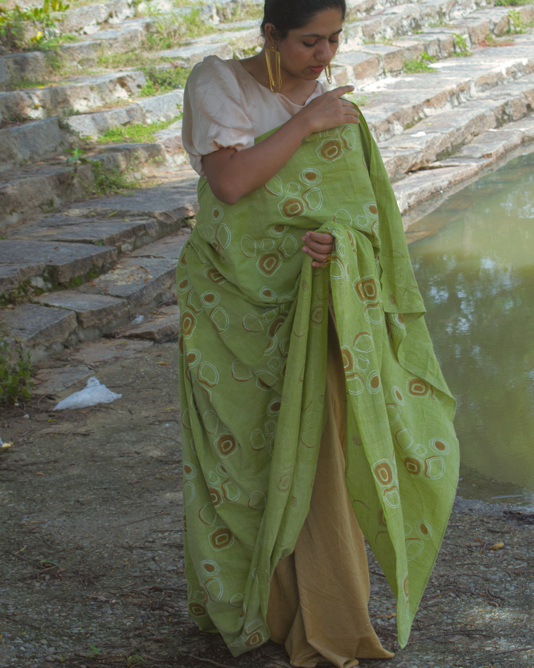 Woman in green and brown saree in front of water body and gold earrings