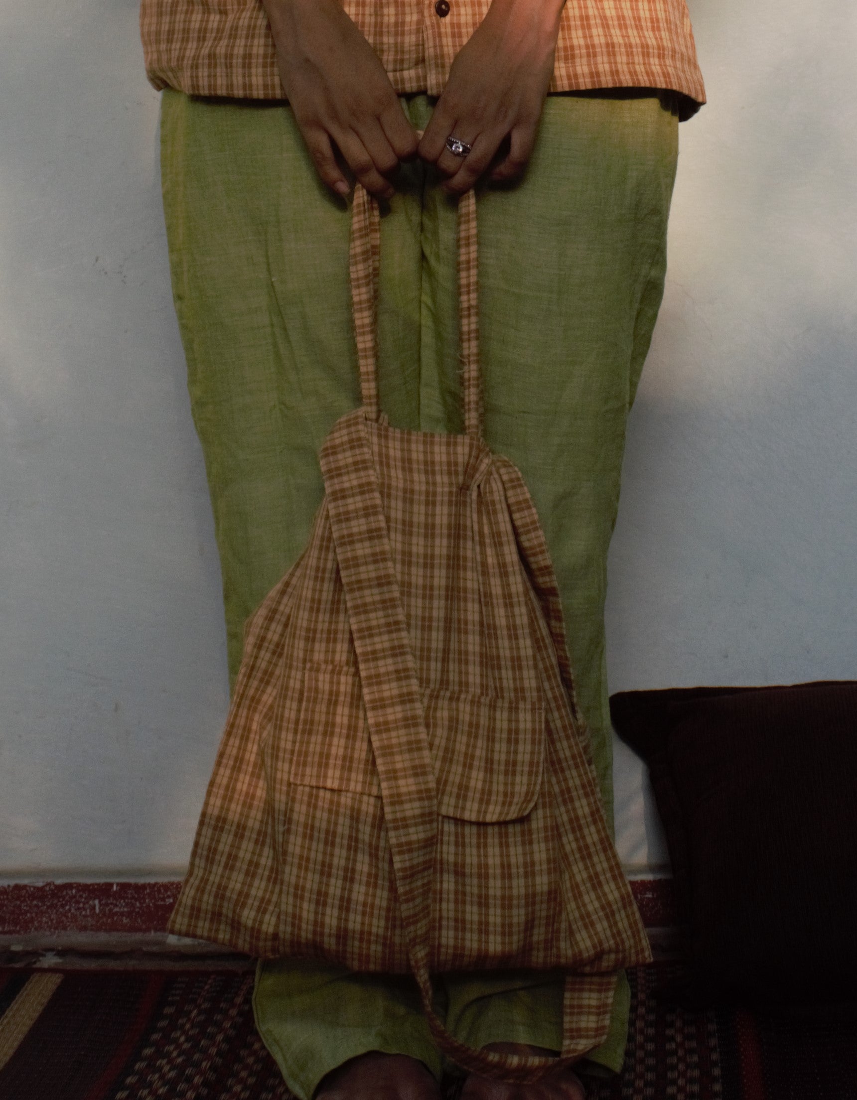 A girl in green pants holding a checks bag which matches her yellow and brown shirt perfectly on a carpet with a cushion 